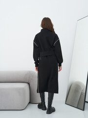 Woman with long brown hair in black dress and knee-high boots stands confidently by modern sofa,...