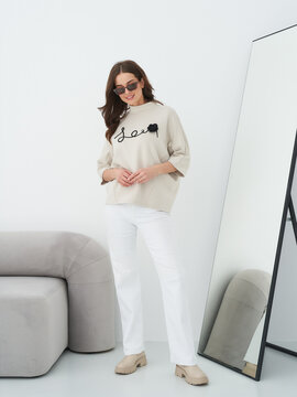 A woman in sunglasses wears a beige top with cursive text, white pants, and tan shoes, standing near a full-length mirror with a gray sofa partly visible. Her top features a black design