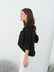 Profile view of a woman with long brown hair wearing a black top and white pants, standing beside a...