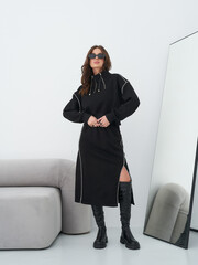 Woman with long brown hair in black dress and knee-high boots stands confidently by modern sofa,...