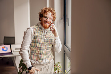 A stylish man with beautifully curly hair is smiling brightly while holding his smartphone in a modern office setting