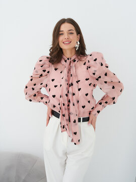 Smiling woman wearing a pink blouse with black heart patterns and white pants, posing with hands on her hips. Her hair is styled in soft waves, and she wears statement earrings