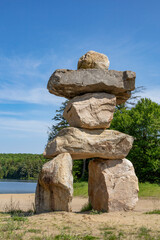 A towering Inukshuk stands proudly by the serene shores of Clear Lake, set against a backdrop of lush greenery and a clear blue sky.