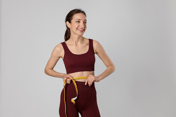Beautiful woman measuring her belly with tape on gray background