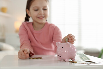 Girl putting her pocket money into piggybank at table indoors, selective focus