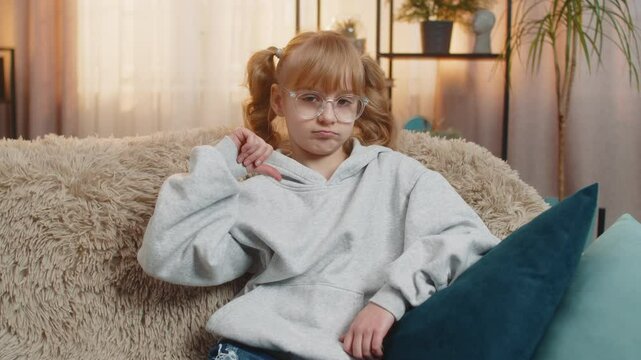 Young teenager girl showing thumbs down dislike with a disappointed expression at home. Caucasian child kid sitting on sofa, expressing dissatisfaction and disagreement through a clear hand gesture.