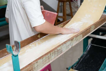 Close-up side view of a food worker operating an industrial dough sheeter, applying flour to a wide...