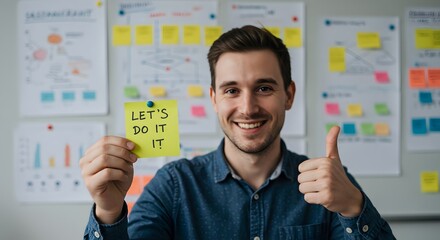 Motivated young businessman holds Lets Do It sticky note smiling giving thumbs up near project planning charts