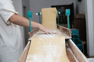 Close-up side view of a food worker operating an industrial dough sheeter, applying flour to a wide sheet of fresh yellow pasta dough