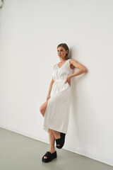 Woman in a white sleeveless dress leans against a wall, with one hand on hip, wearing black...