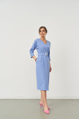 Woman wearing a knee-length light blue dress with a matching belt and pink high heels. She stands...