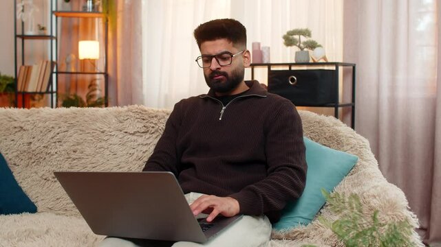 Indian man at home types on laptop with focused expression, preparing for upcoming online exam with full attention. Arabian guy on sofa concentrates on studying and memorizing materials for success