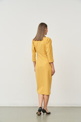 A woman with straight brown hair stands facing away, wearing a mid-sleeve, knee-length yellow dress...