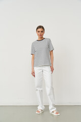 Casual fashion model in striped shirt and white pants poses confidently. Short-sleeved top, relaxed fit trousers, and white sandals. Minimalist style with natural light from the left