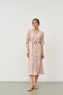A woman with light hair wears a knee-length light pink dress featuring short sleeves, buttoned front, and a belt with a circular buckle. She stands confidently with neutral expression
