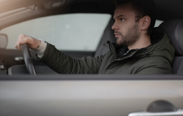 A serious young man in a green jacket is seated in a car, holding the steering wheel with focus....