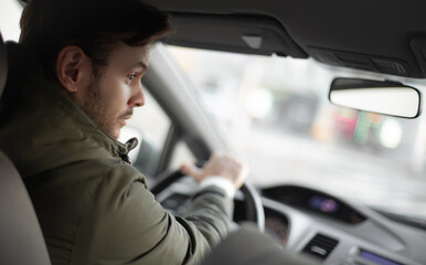Young man driving a car, looking alert and focused while turning the steering wheel. He wears a...