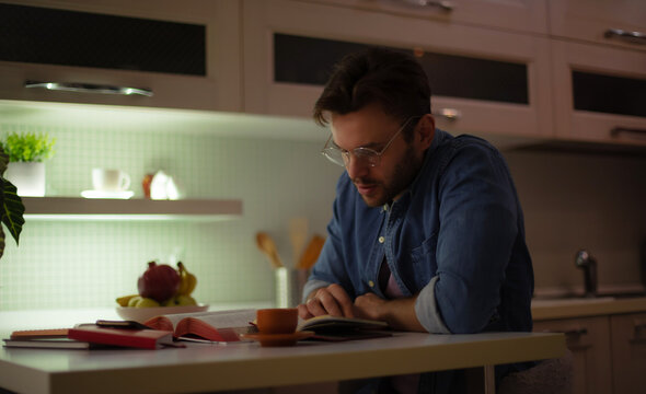 A focused young man in glasses reads a book at a kitchen table at night, surrounded by open books, a cup of coffee, and warm ambient lighting, creating a cozy and studious atmosphere.