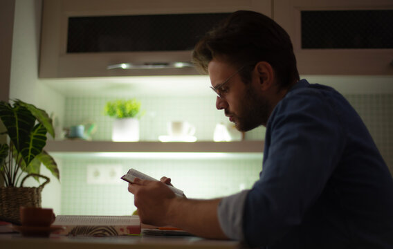Young man with glasses sitting at a kitchen table, deeply focused on reading a book, surrounded by notebooks, a coffee cup, and soft lighting in a cozy modern interior.