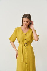 Woman in yellow button-down dress with belt, short sleeves, standing against neutral backdrop. She...