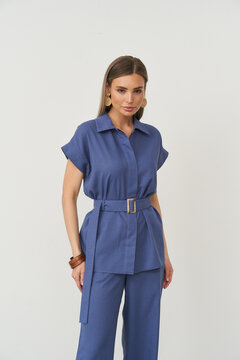 Elegant woman with long hair wearing blue belted shirt and matching trousers, accessorized with large hoop earrings and a wooden bracelet, poses confidently with relaxed expression