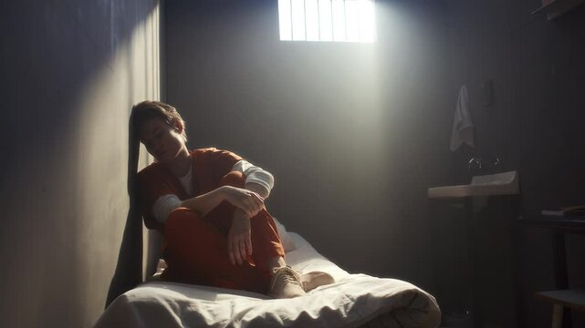 Depressed female inmate, deep in thought on bed, wearing orange uniform, resting her head against wall while sitting during lonely day in prison cell. Zoom shot