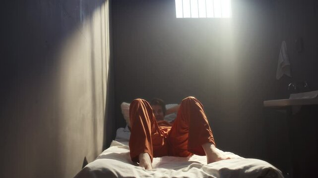 Young female inmate in orange uniform performing sit-ups on prison bed during fitness routine in jail. Zoom-in shot