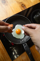 Close-up of hands holding the cracked eggshell just above a sunny side up egg frying in a black pan, highlighting the fresh cooking process and tactile kitchen moment.

