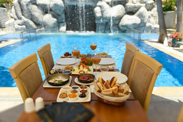 Elegant hotel breakfast setup on wooden table with salad, cheese, olives, jams, nuts, wine glasses, and four wooden chairs near a pool