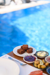 A wooden table set with sesame cookies, olives, butter, jam, tomato, and a fork, with a serene poolside view at a luxury hotel
