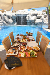 Wooden table set for breakfast with juices, salad, cheese, olives, fruits, and jams. Pool and wooden chairs create a relaxing vacation atmosphere