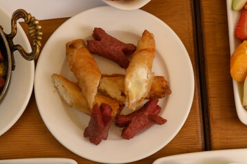 A wooden table seen from above featuring a plate with fried sausages and crispy cheese sticks, part of a hotel breakfast scene