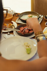 Hotel breakfast scene with a plate of fried eggs and sausages. A woman uses a fork to take a sausage from the serving plate to her own. Cozy and relaxed atmosphere