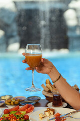 A wooden table set for breakfast at a hotel with salad, olives, tomatoes, and cheese, while a woman’s hand holds a glass of juice. A serene pool is visible in the background