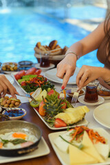 A hotel breakfast on a wooden table with fruits, olives, salad, and cheese as three women use forks to pick fruits with a pool view in the background