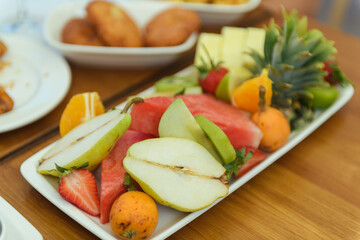 A vibrant mix of fresh fruits including pineapple, strawberry, watermelon, pear, and persimmon displayed on a large plate atop a rustic wooden table in natural light