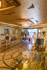 Opulent hotel lobby in baroque style with golden accents, blue velvet furniture, marble surfaces, lavish chandeliers, and three female guests arriving with luggage