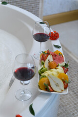 Elegant hotel jacuzzi filled with foam and roses, with wine glasses and fresh fruits on the edge