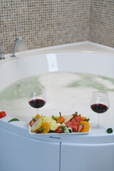 Elegant hotel jacuzzi filled with foam and roses, with wine glasses and fresh fruits on the edge