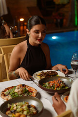 Attractive brunette woman dining at hotel restaurant table filled with various meat dishes and salads, pool visible in background