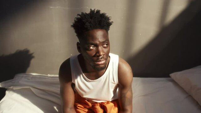 Young Black prisoner sitting on bed, looking at sunlight streaming through barred window and posing on camera in jail cell. Video portrait