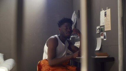 Young Black inmate sitting at desk, looking upwards thoughtfully and writing letter in notepad during day in solitary prison cell - Powered by Adobe