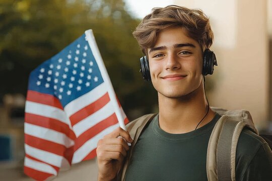 Smiling young man wearing headphones and a backpack holding an american flag outdoors with a blurred natural background