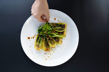 A hand adds garnishing to freshly prepared bok choy on a white plate in a vibrant kitchen setting.