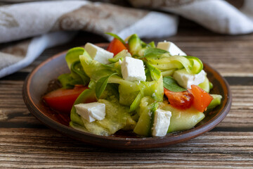 Vegetable salad with raw pickled zucchini, feta cheese and fresh tomatoes on clay plate with cloth napkin on wooden table, side view, close up. Healthy diet.