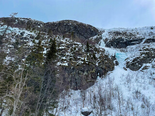  Snow-covered rocky mountain with patches of ice and sparse trees under a cloudy winter sky. The...