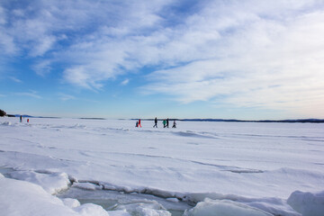  A wide frozen lake under a bright blue sky with scattered clouds. People in colorful clothes are...