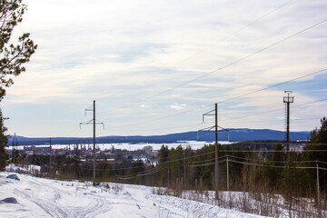  A snow-covered clearing with power lines stretching across the winter landscape. Tall pylons line up along the edge of a pine forest under a pale blue sky, creating a mix of nature and infrastructure