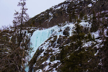  Frozen waterfall cascading down a rocky mountain slope covered with snow and ice. Winter...