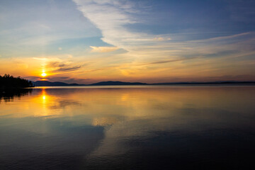 Obraz premium Stunning sunset over a calm lake with golden reflections, mountains on the horizon, and forest silhouette. Perfect for travel, nature backgrounds, peaceful landscapes, eco tourism, and inspiration.